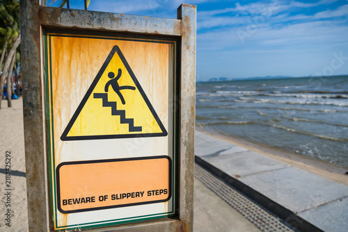 Beware of Slippery Steps Warning Sign on Beach: A cautionary sign depicting a falling figure on stairs, warning beachgoers of slippery steps, stands by the ocean under a clear blue sky.