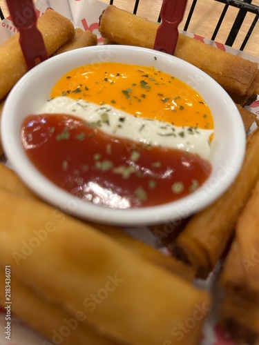 Fotografie The image shows a small, round white bowl filled with three distinct dipping sauces arranged in stripes, served alongside crispy golden spring rolls or lumpiang shanghai