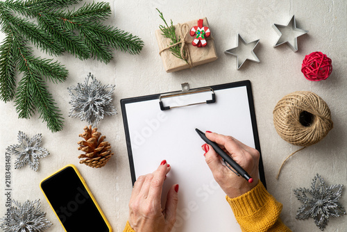 Hands writing on blank paper surrounded by Christmas decorations, gift, smartphone and fir branches. Christmas organization
