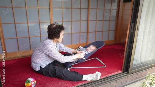 A seated person leans forward with both hands toward an infant reclining in a baby bouncer by sliding doors.