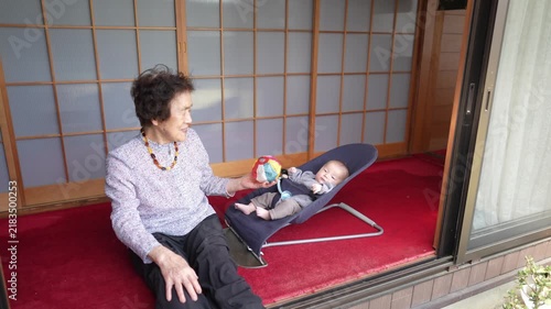 A person extends a colorful patterned ball toward an infant reclining in a baby bouncer by sliding doors.