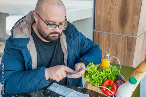 Concerned man check grocery receipt while sit in supermarket with shopping bag filled with food products. Daily expenses, smart budgeting, saving money, price awareness, personal finance