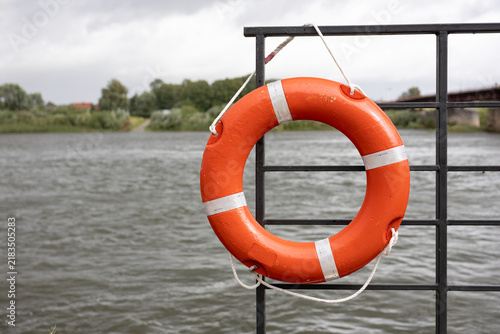 Cuadro en lienzo Orange lifebuoy on metal railing by water on cloudy day