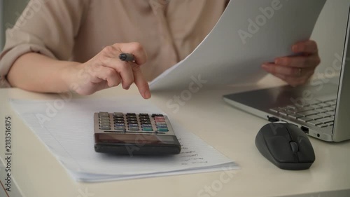Footage of a woman calculating expenses and reviewing paperwork beside a laptop. Business footage illustrating budgeting, accounting, bookkeeping, financial analysis and office administration.
