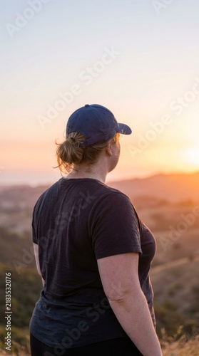 Woman in cap looking at sunset over rolling hills, contemplation in nature during golden hour.