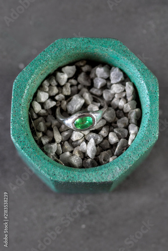 A green gemstone ring rests on gray pebbles inside a green hexagonal pot. The scene highlights jewelry and decorative elements.