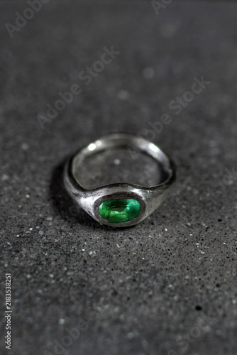 A silver ring with a green gemstone sits on a dark surface. The ring has a simple design and a polished finish, highlighting the vibrant color of the stone.