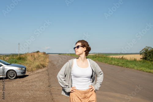 Obraz na płótnie woman standing with hands on hips beside a parked car and empty country road, ta
