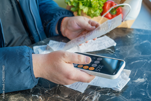 Shopper uses smartphone calculator while reviewing grocery receipts beside bag of everyday food. Rising shopping expenses, inflation, cost of living pressure, household financial budgeting
