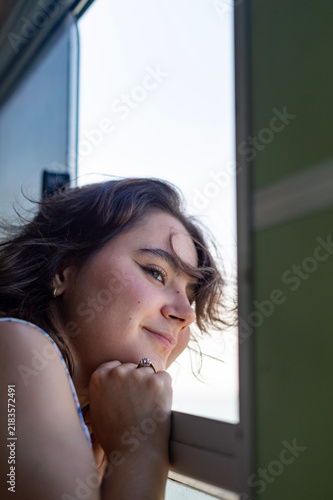 A young woman leans her chin on her hand while looking out an open window in Venice, Italy. Bright natural daylight illuminates her face in a vertical side profile view.