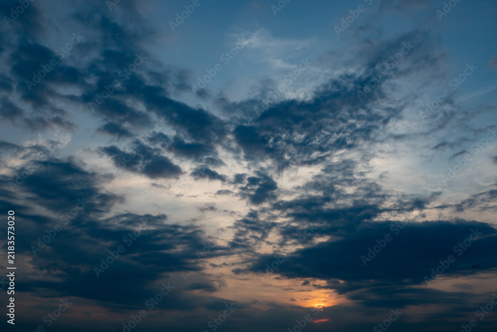 Obraz premium A horizontal sunset sky with dark blue-grey clouds and a warm orange glow breaking through at the bottom right. Moody atmosphere with dramatic cloud layers and copy space.