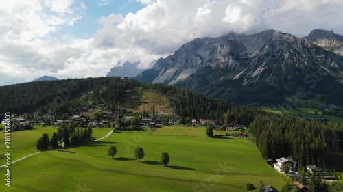 Aerial drone view of the Dachstein mountain range and Ramsau am Dachstein in Austria. Slow flight over green alpine meadows, traditional mountain houses and forests in warm summer sunset light.