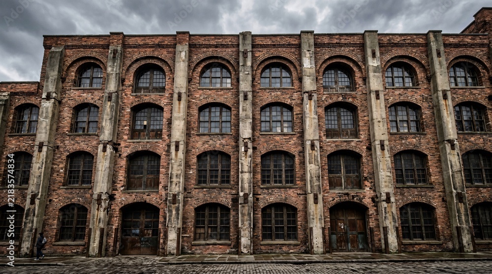 Obraz premium Historic industrial facade of weathered red and brown bricks, arched window frames with dark mullions, concrete pillars, overcast sky