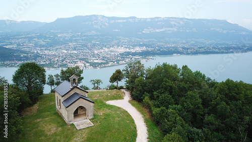 Aerial view of the Notre-Dame-de-l'Etoile chapel on a hill overlooking Lac du Bourget and lush forest in La Chapelle-du-Mont-du-Chat, Auvergne-Rhone-Alpes, France.