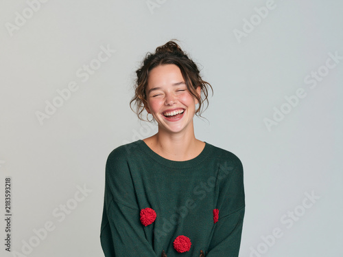 Happy young woman with hair bun wearing a green Christmas sweater laughing sincerely with closed eyes on a light background