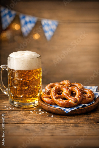Golden beer in a glass with frothy head. Baked pretzels sit on a wooden plate. Blue and white bunting hangs above the table. Cozy wooden background with soft bokeh lights. Perfect for festive, rustic