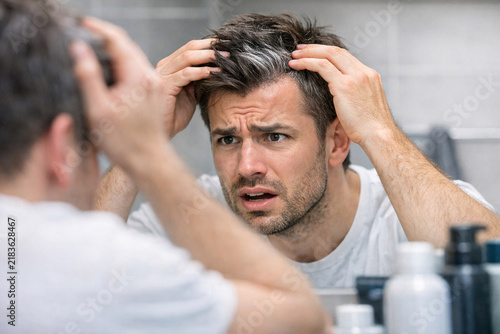 A man looking at his reflection in a mirror, shaving his beard and mustache. He has a thoughtful expression as he grooms his facial hair.