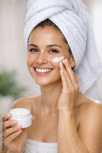 Smiling woman with towel wrapped around her, applying a skincare product with both hands.