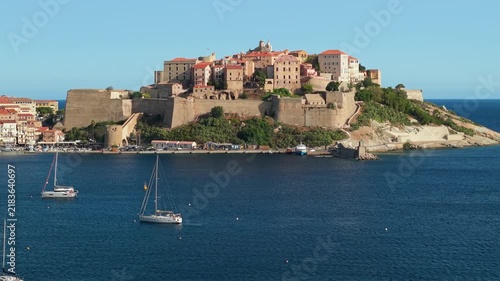 Aerial 70mm telephoto drone view of Calvi Citadel with sailboats in Corsica France. Luxury sailing yachts anchored near historic peninsula 4K