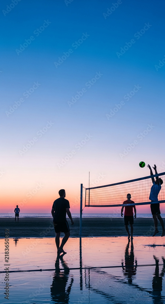 Fototapeta premium Silhouettes of people playing volleyball on a wet beach at sunset, symbolizing recreation, sport, and peaceful evenings