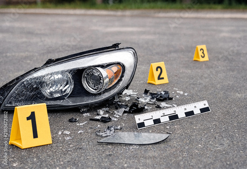 Shattered car headlight on asphalt with yellow evidence markers and a scale.