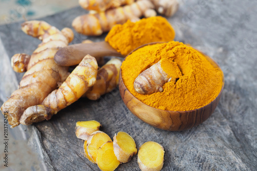 Fresh turmeric roots with sliced pieces and turmeric powder in wooden bowl on rustic wooden background.