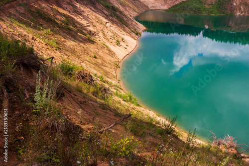 Stunning turquoise water fills a deep sand quarry lake surrounded by rugged earthy slopes and wild greenery under a clear sky in this peaceful wilderness landscape