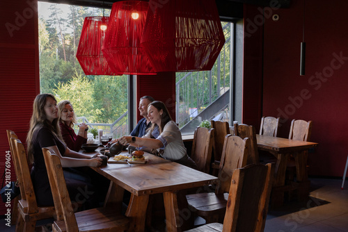 Two generations of a family dining in a wooden hall under red woven lamps