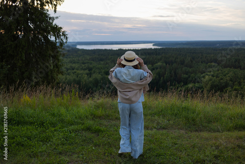 Rear view of a woman holding her straw hat above a green valley with a river at sunset