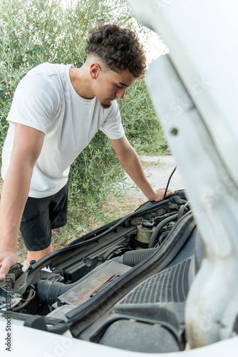 Fotografie Young man standing by open hood, looking at engine of broken down car on rural r