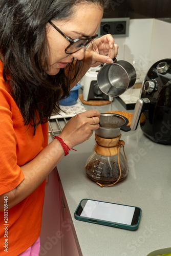 Woman in glasses carefully pouring hot water into a coffee filter, brewing pour over coffee in her kitchen with a smartphone nearby