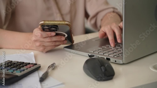 A woman tapping a smartphone and working on a laptop at an office desk with calculator and paperwork. Close up business footage representing multitasking, remote work, and digital communication. 