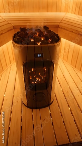 Sauna heater with glowing rocks and digital display emits steam inside a wooden sauna, showcasing the warm ambiance and natural wood textures of the interior space