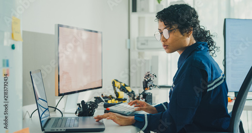 Young African American female robotics engineer coding AI software for robotic arm in laboratory. Embedded system, firmware testing, machine learning, industrial automation, future technology concept.