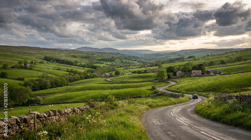 Winding country road through green rolling hills and rural landscape under dramatic clouds.