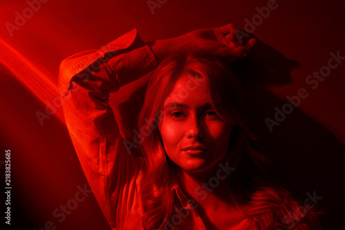 Moody portrait of a young woman bathed in red light, posing against a wall with dramatic shadows and a contemplative, sensual expression for fashion, editorial, and cinematic use.