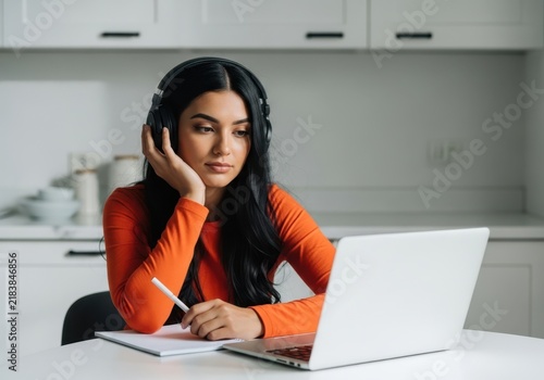 Young woman in orange shirt wearing headphones studying with laptop in kitchen