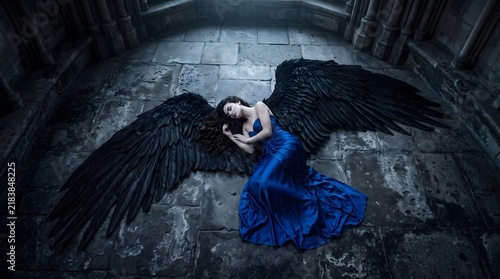 A beautiful woman with large black angel wings wearing a flowing blue dress lying on a stone floor