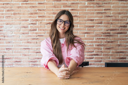 Фотография Young woman wearing eyeglasses sitting at a wooden table in front of a brick wal