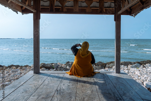 Принти на полотні Woman wearing yellow hijab sits inside wooden pavilion by ocean for peaceful relaxation