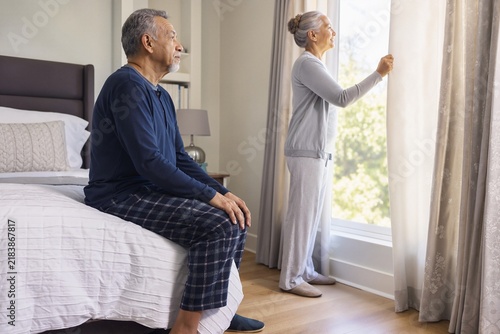 Seniors enjoying morning light with elderly man sitting on bed and woman opening curtains. Morning moment brings warmth and hope for elderly couple as they embrace new day together.