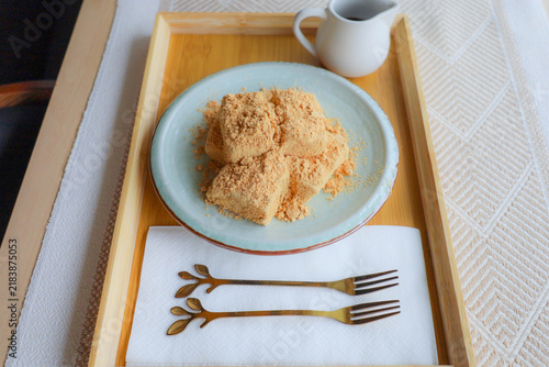Traditional Japanese warabi mochi dusted with kinako soybean powder served on a ceramic plate on wooden tray with kuromitsu syrup and brass forks.