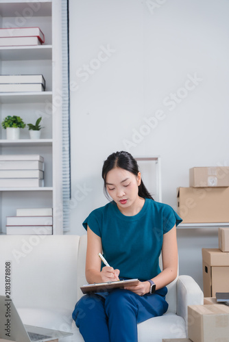 Fotografie Focused Entrepreneur: A young Asian woman sits on a sofa, diligently writing in a notebook with a stylus, surrounded by boxes, implying a small business owner managing inventory or orders