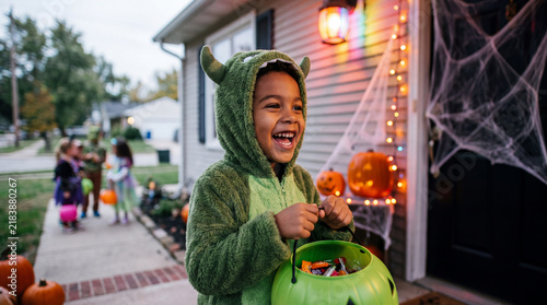 Halloween monster child trick or treating with green pumpkin bucket at doorway, a silly playful autumn celebration photo for social media and banners.