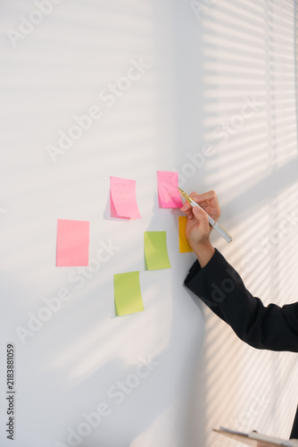Strategy Session: A hand holding a pen writes on a sticky note affixed to a wall, surrounded by colorful notes, in a bright, sunlit room.