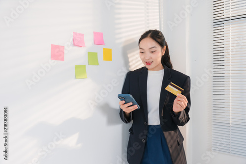 Fotografía Online Shopping Transaction: A young Asian woman in a professional setting holds a smartphone and a credit card, engaged in an online transaction