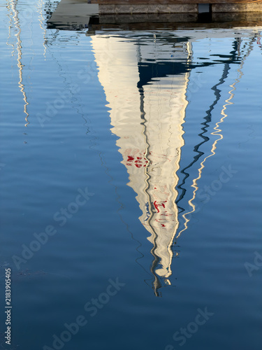 Inverted mirror-like reflection of a sailboat's white hull and dark cabin with red lettering, rippling in calm blue water