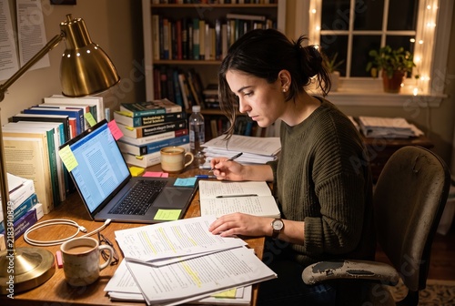 A student is working late at night at a desk covered with books, notes, and a laptop. The room has bookshelves in the background and warm light from a lamp