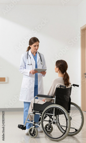 woman white coat holding tablet stands hospital hallway patient wheelchair looking medical