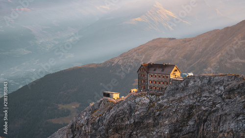 Julius Payer Hut stands on a rocky ledge in the Italian Alps near Ortler Mountain. The sun sets behind the mountains, casting shadows and creating a scenic landscape.